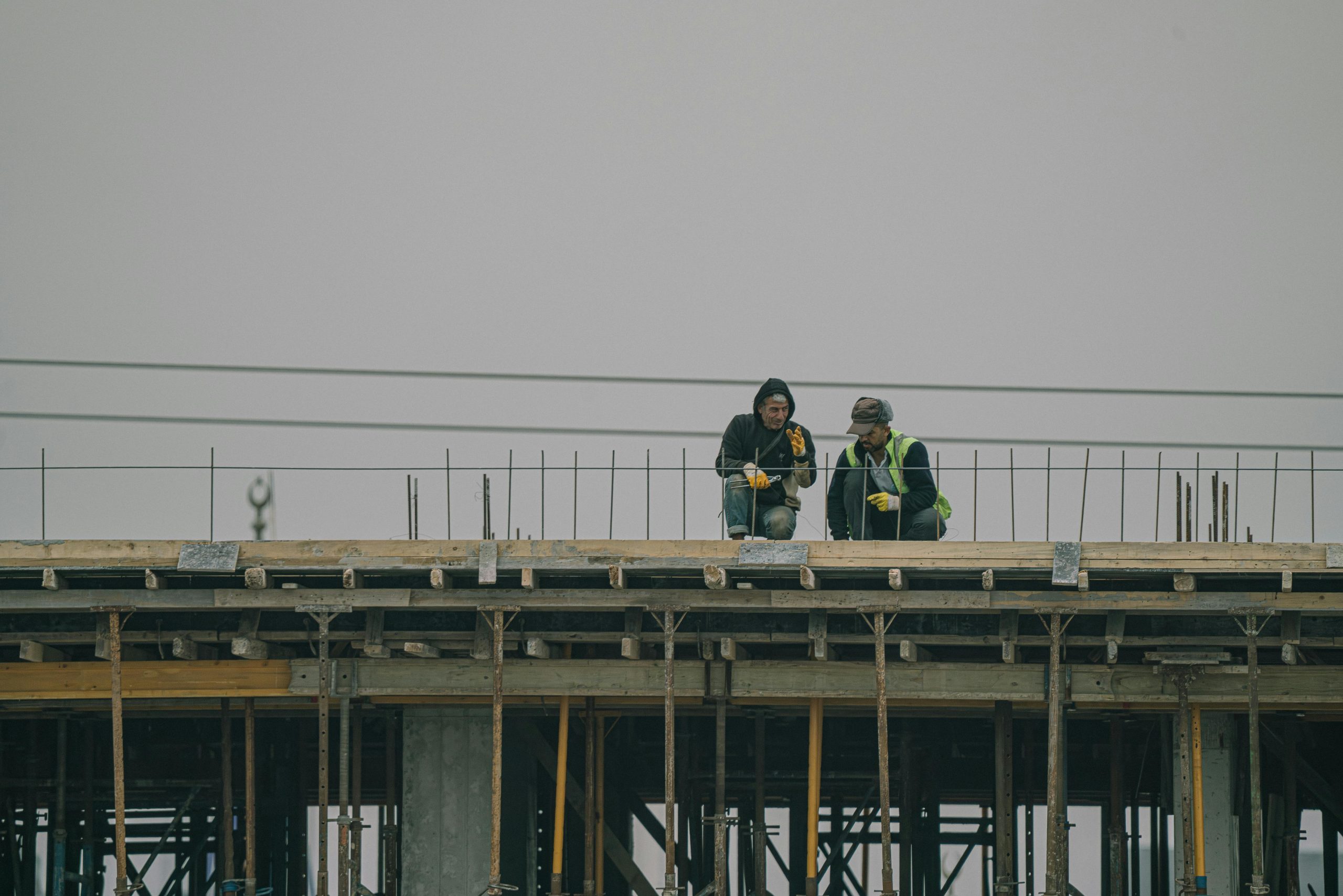 Home Two construction workers on a building site in Denizli, Türkiye, working under overcast skies.